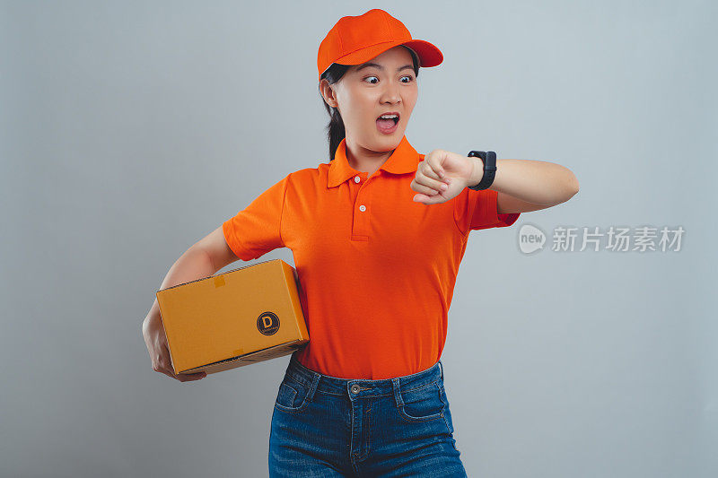 亚洲女人穿着统一的polo t恤，拿着盒子查看时间，因为给客户送包裹迟到而震惊，孤立地站在白色的背景上。